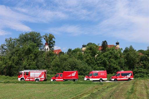 Vier Feuerwehrautos auf einem Weg, davor grüne Wiese, dahinter grüne Bäume, blauer Himmel